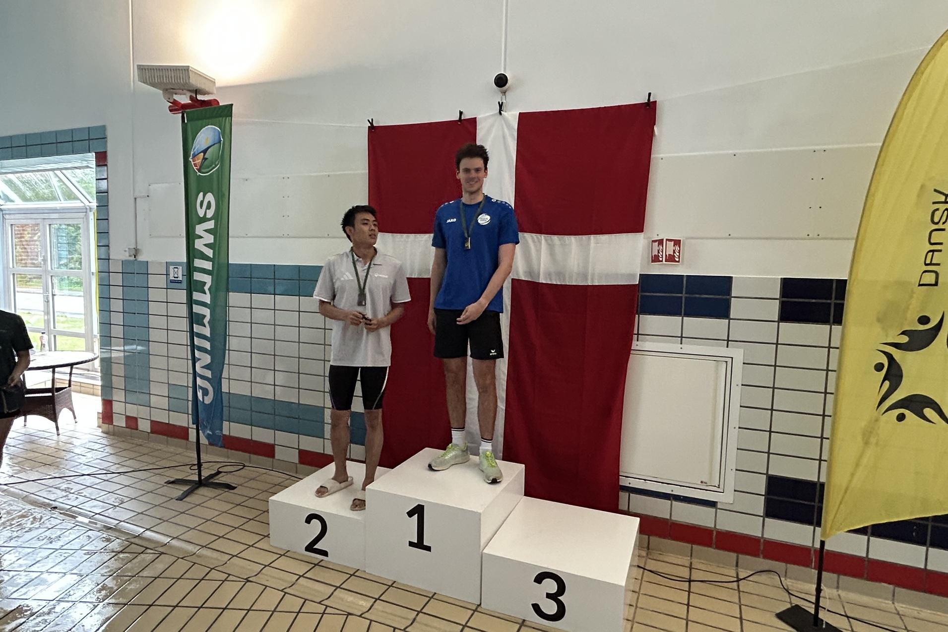 Zwei Schwimmer stehen auf dem Siegertreppchen, der erste in blauer Schwimmbekleidung, hinter ihnen eine dänische Flagge.
